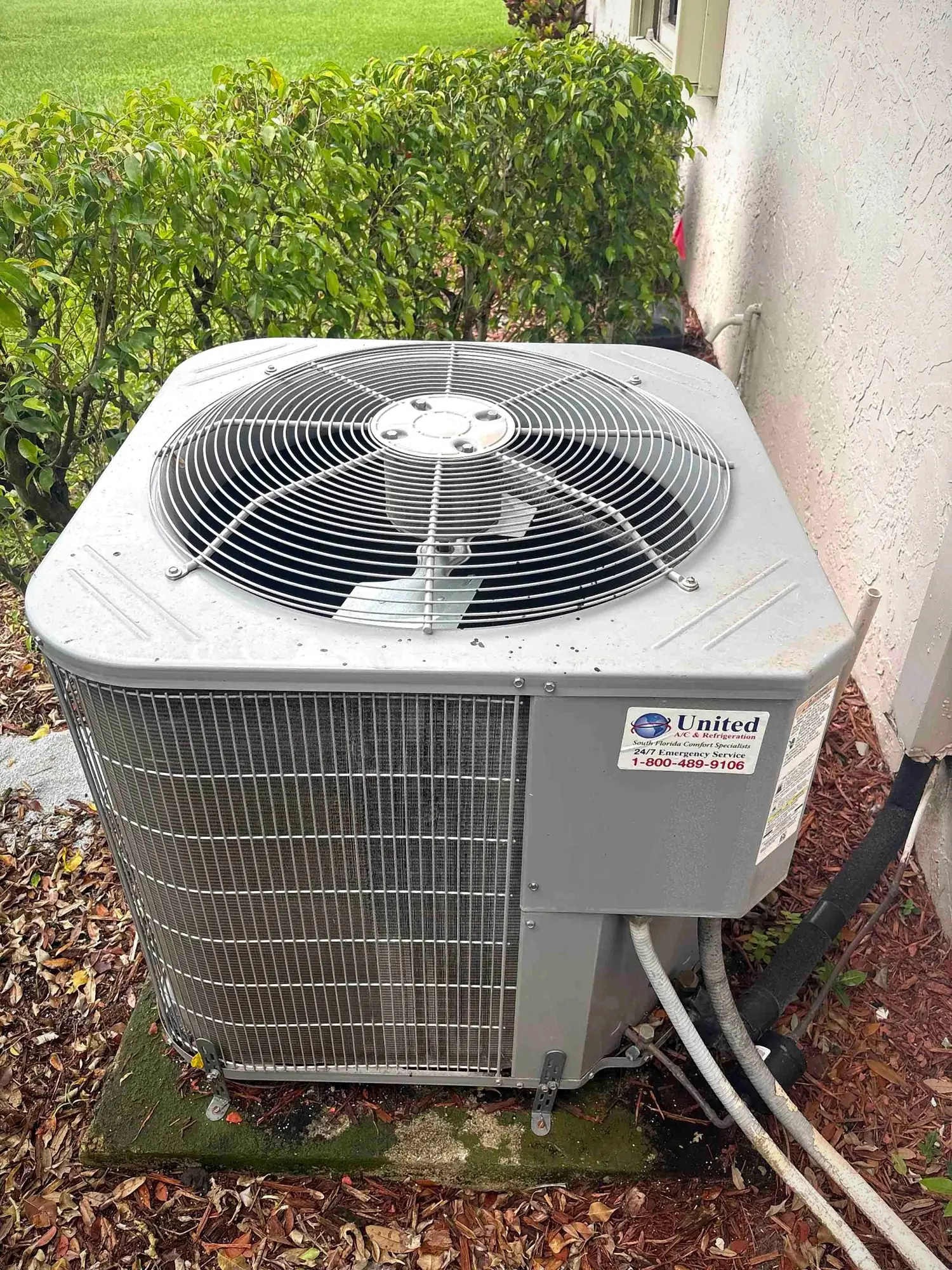 Residential air conditioning condenser on a concrete pad beside a hedge and stucco wall