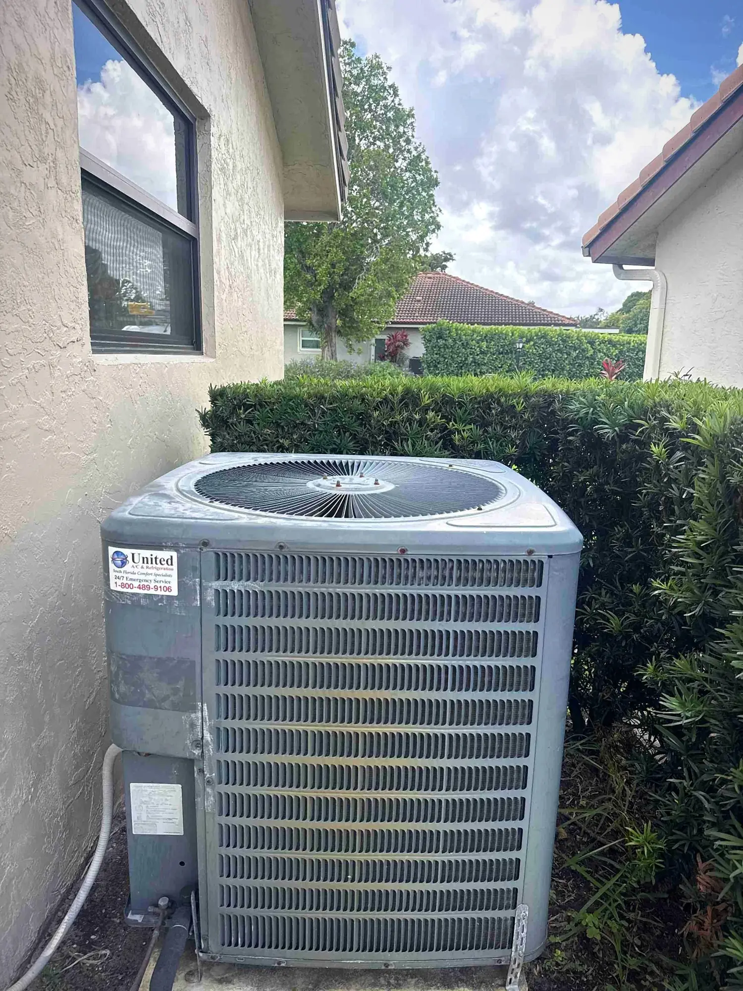Residential air conditioning condenser on a slab beside a stucco home and hedge