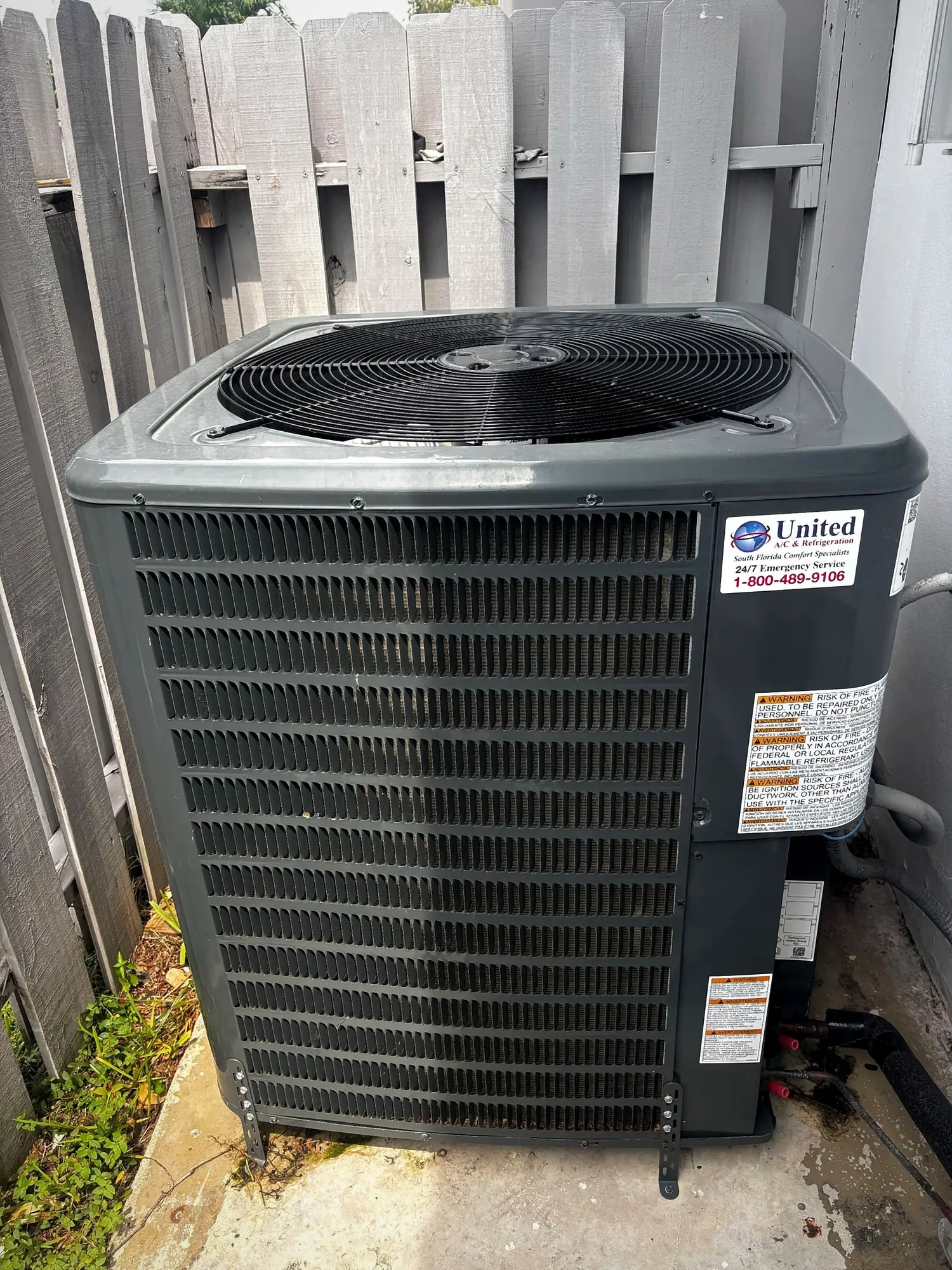 Outdoor condensing unit on a concrete pad beside a wood fence with refrigerant line connections